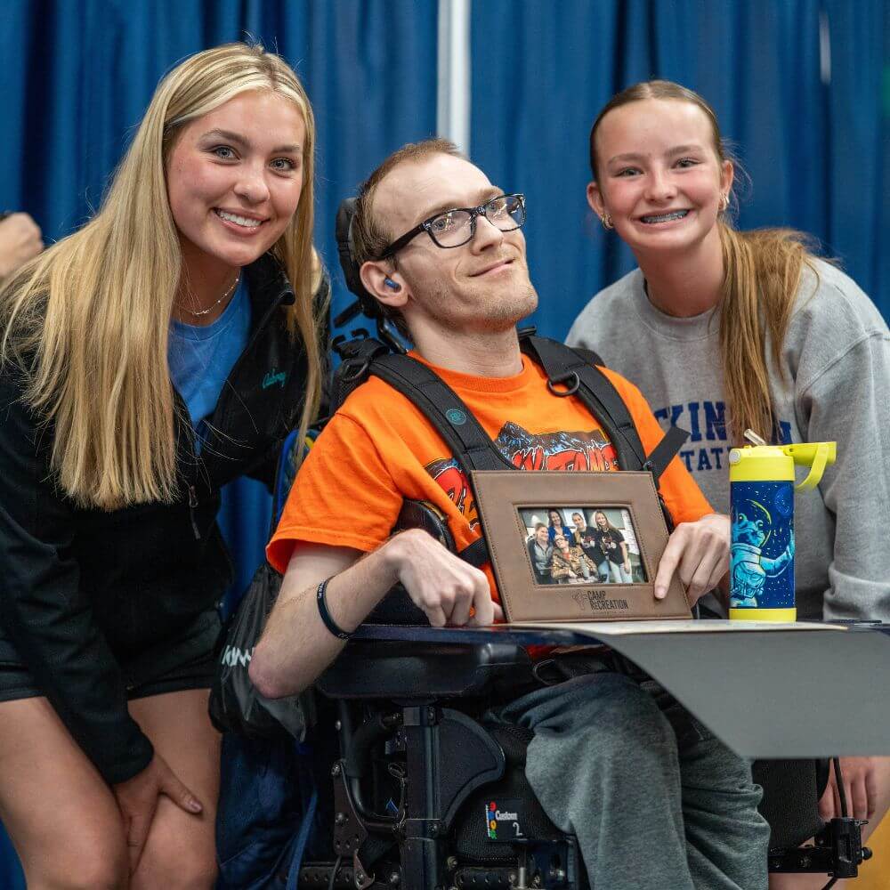 Male camper in wheelchair holding photograph between two female volunteers