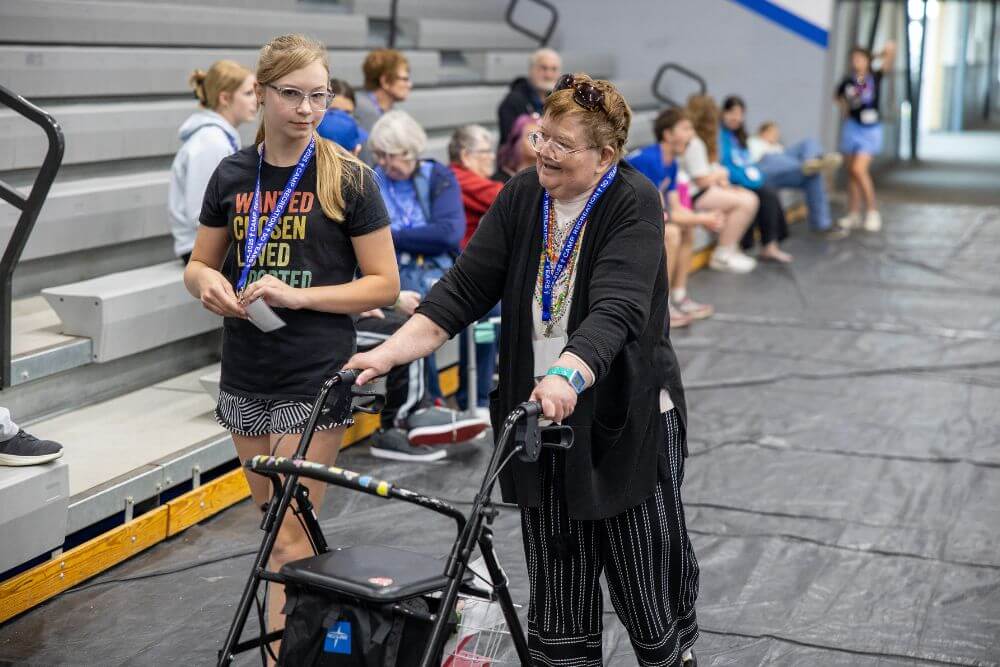 Volunteer walking with camper with walker in front of bleachers at Camp ReCreation
